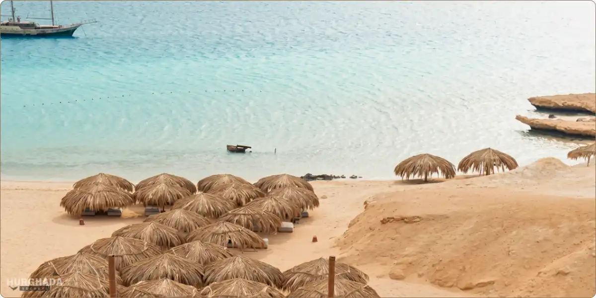 Aerial view of many traditional straw umbrellas on the sandy beach of Ozirea Island in Hurghada, with clear turquoise water and a boat in the background.