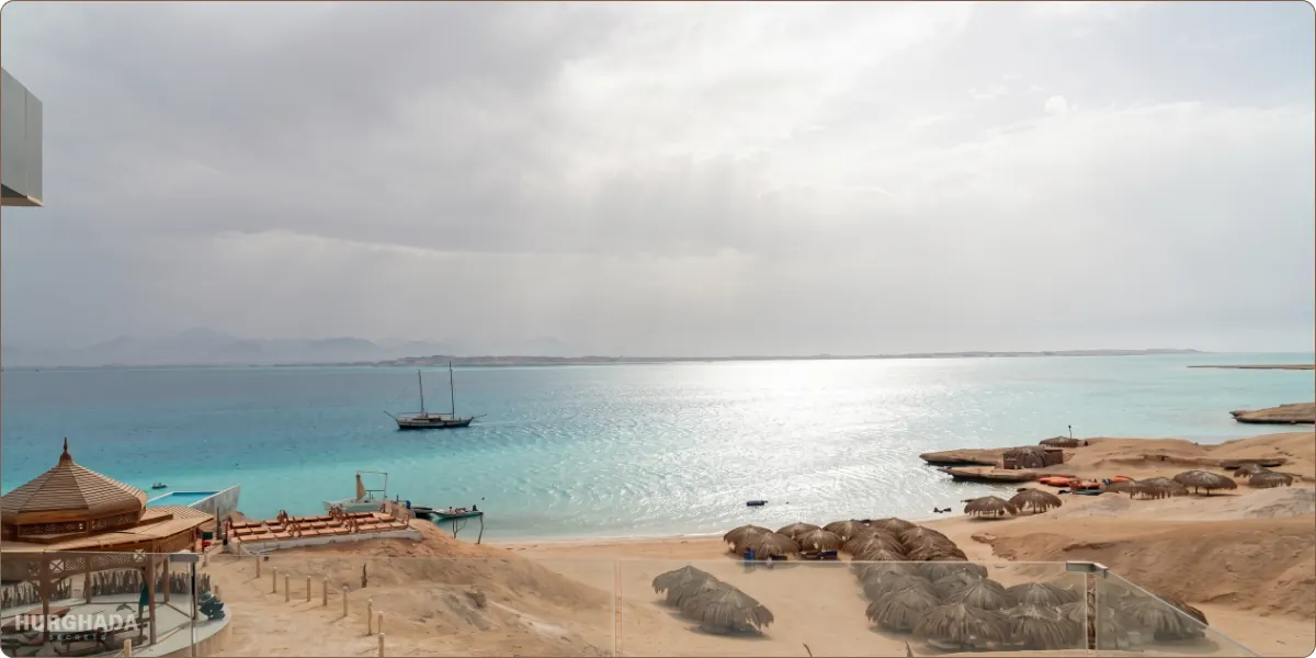Panoramic view of Ozirea Island beach in Hurghada featuring turquoise Red Sea waters, a traditional sailing boat, and straw umbrellas on the sandy shore.