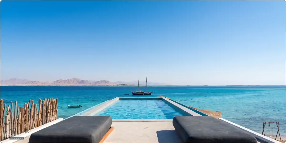 Luxury infinity pool on Ozirea Island in Hurghada, offering a stunning view of the turquoise Red Sea with a traditional boat and distant mountains under a clear sky.