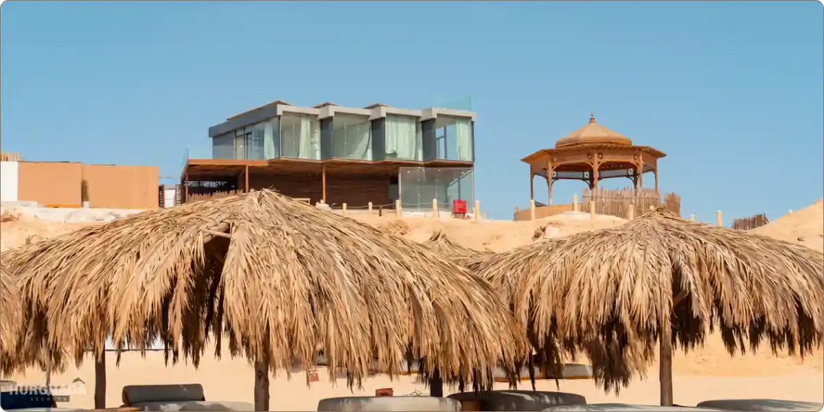 Modern glass villa and a traditional wooden gazebo on the sandy dunes of Ozirea Island in Hurghada, viewed from behind straw beach umbrellas.
