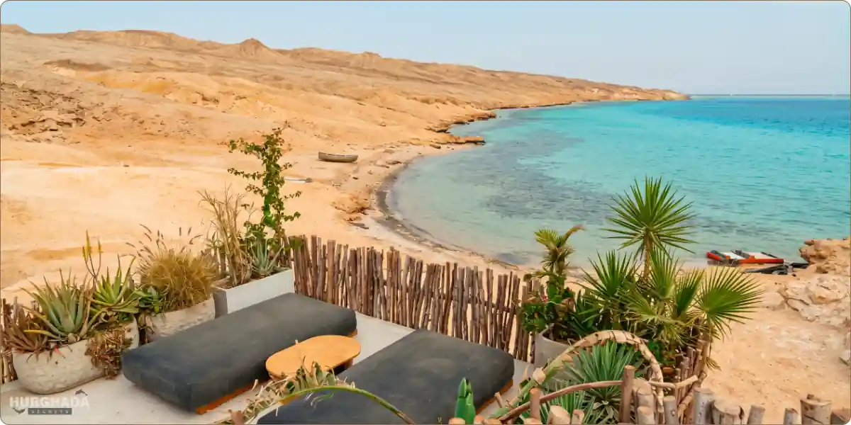 Relaxing beach lounge area on Ozirea Island in Hurghada, overlooking a crystal-clear blue bay surrounded by natural desert hills and greenery.
